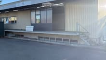 Modern industrial-style building with a metal facade, large windows, and a covered entrance area. Features a concrete staircase leading to the entrance and a small bench near the entrance.
