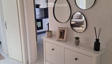 Hallway with white tiled floor, light-colored walls, and a small console table with decorative items, mirrors, and a view into a kitchen and bedroom area.