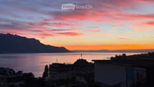 View from a balcony or terrace overlooking a lake and mountains at sunset, with city buildings in the foreground.