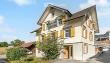 Two-story house with light-colored walls, yellow shutters, and a brown roof. The house has multiple windows and a central entrance with a wooden door. There is a small garden area in front of the house, and a sign on the wall indicates it is for sale.