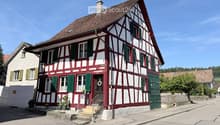 Traditional half-timbered house with red and white exterior, green shutters, and a small garden area with potted plants near the entrance.