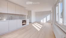 Modern kitchen with white cabinets, light wooden flooring, and large windows allowing natural light to fill the space. Features include a sink, faucet, and built-in oven.