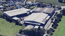 Aerial view of a residential and industrial area with multiple buildings, including houses, warehouses, and a large industrial complex with a prominent roof structure. The area is surrounded by green fields and trees.
