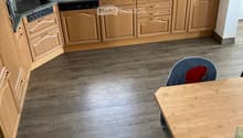 The kitchen features light wooden cabinets, a granite countertop, and a wooden dining table with a single chair. The floor is made of wood, and the sink area includes a faucet and various small items.