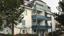 A multi-story residential building with light blue shutters, balconies, and a well-maintained lawn.
