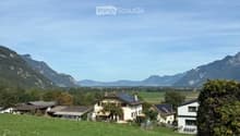 A peaceful rural landscape with a cluster of houses, green fields, and distant mountains under a clear blue sky.