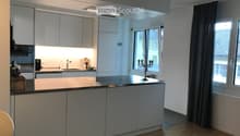 Modern kitchen with white cabinetry, a central island, and a sink area. Features include a candle holder on the island, a window with dark curtains, and built-in lighting.