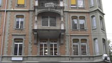 Four-story brick building with arched windows, balconies, and decorative stonework. Features a central entrance with large glass doors and a small green area in front.