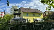 Two-story yellow house with a brown tiled roof, multiple windows, and a balcony on the upper level. The house is surrounded by greenery and a metal fence.