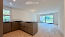 Modern kitchen with light wooden flooring, grey cabinets, and a black countertop. Features a large window and a sliding glass door leading to an outdoor area.