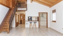 Open-plan living and dining area with a wooden staircase leading to an upper level, a kitchen area with cabinets and a range hood, and two stools at a kitchen island.