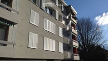 Four-story residential building with grey facade, white shutters, and multiple balconies with green awnings.
