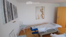 A small, well-lit room with a treatment table, two chairs, and artwork on the walls.