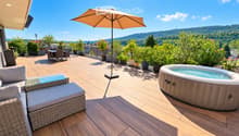 Wooden deck with a sofa, glass coffee table, dining table with chairs, umbrella, and a hot tub, surrounded by potted plants and offering a scenic view of the valley.