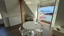 Bright attic room with wooden floors, white furniture, and a large window offering a scenic view of the lake and mountains. The room features a round white table with two chairs, a desk with a chair, and a white cabinet.