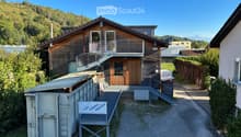 Two-story wooden house with a balcony, staircase, and a small shed with a ladder. The house is surrounded by greenery and has a clear view of mountains in the background.