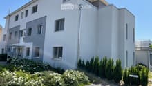 Modern two-story house with white and grey exterior, multiple windows, and a well-maintained garden with shrubs and flowers.