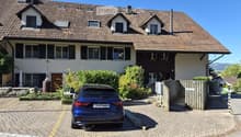 Two-story house with a tiled roof, multiple windows, and a garage. A blue car is parked in the driveway.