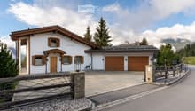 A two-story house with a white exterior, wooden garage doors, and a stone driveway. The house features a traditional alpine design with wooden shutters and a sloped roof. There is a decorative wooden fence and a small statue near the entrance.