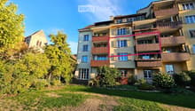 Multi-story residential building with multiple balconies, surrounded by greenery and a lawn.