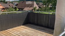 Wooden deck with metal railing, overlooking a residential area with houses and trees under a partly cloudy sky.