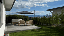 A modern outdoor patio with a wooden deck, white sofa, two wicker chairs, and a large umbrella, surrounded by greenery and a scenic mountain view.