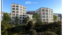 Modern multi-story residential building with white facade, large windows, and surrounding greenery.