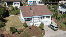 Aerial view of a modern two-story house with a tiled roof, white exterior, and a garage. The house features a raised ground floor, a balcony, and is surrounded by greenery and neighboring houses.