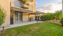 Well-maintained garden with a neatly trimmed lawn, a patio area with a pergola, a barbecue, and a small storage shed. The house features a balcony with plants and flowers, and the exterior is made of light-colored brick.
