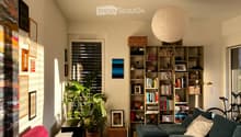 Living room with two sofas, a round red stool, a bookshelf, a coffee table, and a large plant near the window. The room features a colorful rug and modern decor.