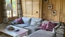 Cozy living room with light wooden walls and ceiling, grey sectional sofa with purple and grey cushions, white coffee table with remote controls and a basket, floor lamp, and decorative picture frames on the wall.