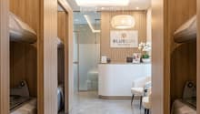 Modern hallway with wooden paneling, recessed lighting, and a reception area visible at the end. Features a sleek, minimalist design with a polished concrete floor and a reception desk with chairs.
