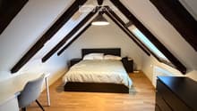 Cozy attic bedroom with wooden floors, slanted ceilings, and large windows. Features a bed with white bedding, a nightstand, a desk with a chair, and a hanging pendant light.