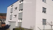 Modern multi-story building with white facade, grey accents, and multiple windows with shutters. Small garden area in front with some plants and a clothesline.