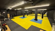 A spacious gym area with a large yellow and blue mat, boxing bags hanging from the ceiling, weights, a bench, and a mirror on the wall.
