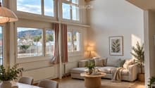 Bright, airy living and dining area with wooden floors, large windows, a grey sectional sofa, a round coffee table, a dining table with chairs, and decorative plants.