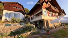 Two-story house with wooden exterior, red shutters, and a balcony. The garden features potted plants, a stone wall, and a patio area with outdoor furniture.
