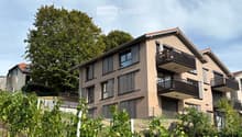 Two-story house with beige facade, multiple windows, balconies, and a garage. Surrounded by greenery and a vineyard in the foreground.
