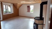 Empty attic room with wooden slanted ceiling, tiled floor, and a fireplace in the corner. Two windows provide natural light.