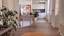 A modern, well-lit hallway leading to a kitchen area. The hallway features a round woven rug on a wooden floor, a small table with a plant and a map, and a chair. The kitchen area has white cabinets and a countertop with various items.