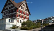 Traditional Swiss-style building with a steeply pitched roof, exposed wooden beams, and red shutters. The structure is two stories with multiple windows and a small balcony on the upper level.