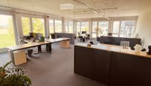 Bright office space with large windows, multiple desks, chairs, and office equipment.
