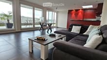 Modern living room with a grey sectional sofa, coffee table, laptop, and large windows leading to a balcony.