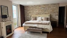 Elegant bedroom with a large bed, fireplace, mirror, and decorative lamp. Features a stone accent wall and large window with curtains.
