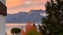 Balcony overlooking a serene lake with distant mountains, surrounded by trees and a parked car.