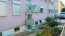 Pink residential building with multiple windows, a small garden area with plants and flowers, and a parked car in the background.