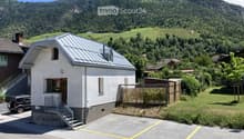 Single-story house with a grey metal roof, white walls, and a small staircase leading to the entrance. The house is situated in a scenic valley with vineyards and mountains in the background.