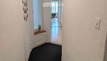 A clean, well-lit hallway with a dark carpeted floor, white walls, and a small window with blinds. There is a coat rack on the left and a small plant on a shelf near the window. The hallway leads to an open area with more rooms visible in the background.