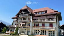A large, multi-story house with a red-tiled roof, white walls, and multiple balconies adorned with striped awnings and flower boxes. The house features a mix of traditional and modern architectural elements, including wooden beams and large windows.