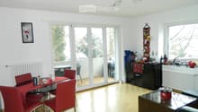 Bright living and dining area with wooden flooring, large windows, and a balcony access. Features a dining table with red chairs, a black cabinet, a sofa, and decorative items on the windowsill.
